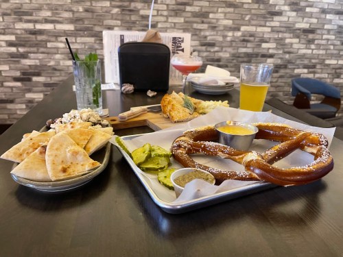 A table with a large pretzel, dipping sauce, pickles, and plates of pita and popcorn, alongside drinks.