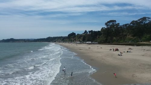 A sandy beach with gentle waves, people enjoying the sun, and cliffs in the background under a blue sky.