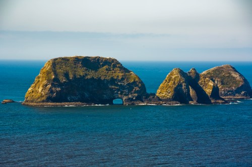 Three rocky islands rise from the ocean, with lush greenery on top and a natural arch in one of the formations.