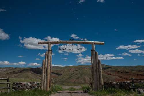 A wooden archway with a sign reading "Lazy J Ranch" against a backdrop of rolling hills and a blue sky with clouds.