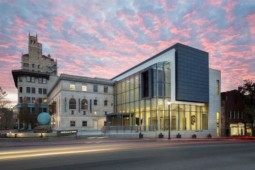 Modern building with glass facade next to a historic structure, set against a colorful sunset sky.