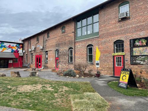 A brick building with red doors and windows, featuring an art gallery sign and colorful murals on the side.