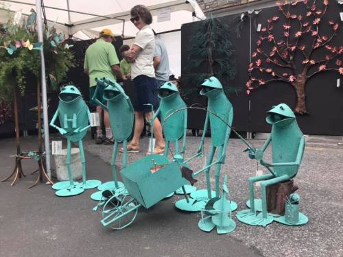 A group of metal frog sculptures in various poses, including fishing, set up at an outdoor market.