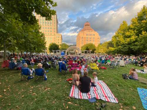 A large crowd enjoys an outdoor concert in a park, seated on blankets and chairs, with buildings in the background.