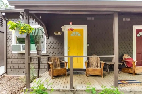 A cozy porch with two wooden chairs, a bright yellow door, and hanging plants on a brown shingle house.