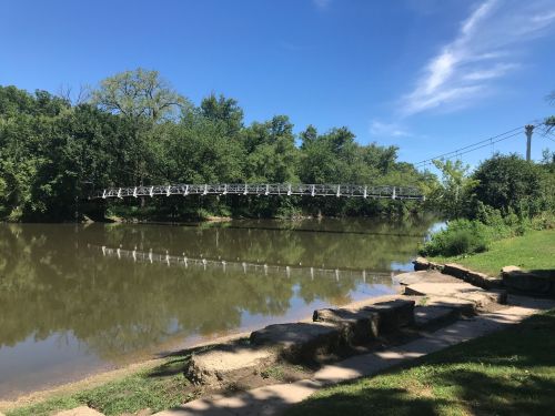 A serene river scene featuring a white pedestrian bridge surrounded by lush greenery and clear blue skies.