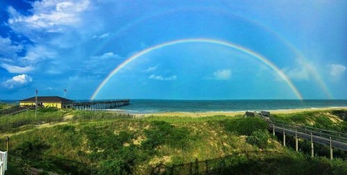 A scenic view of a beach with a double rainbow arching over the ocean and a pier in the background under a blue sky.