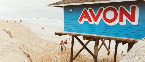 A beach scene featuring a blue building with "AVON" signage, sandy shore, and people walking along the water.