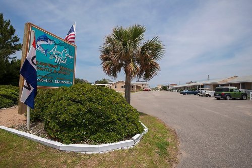 Sign for a beachfront motel with palm trees and flags, leading to a parking area and buildings in the background.