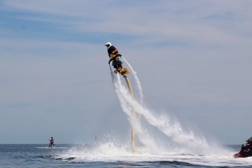 A person using a jetpack above the water, with spray and waves around them on a sunny day.