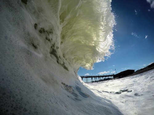 A close-up view of a crashing wave with a pier in the background under a bright blue sky.