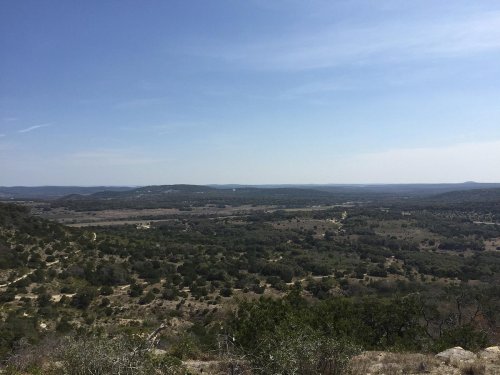 A panoramic view of rolling hills and valleys under a clear blue sky, showcasing lush greenery and distant mountains.