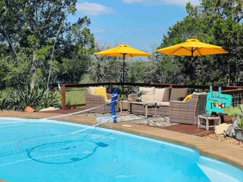 A sunny poolside area with yellow umbrellas, lounge chairs, and a welcome sign, surrounded by greenery.