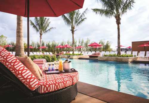 Lounge chair by a pool with drinks and an ice bucket, surrounded by palm trees and red umbrellas.