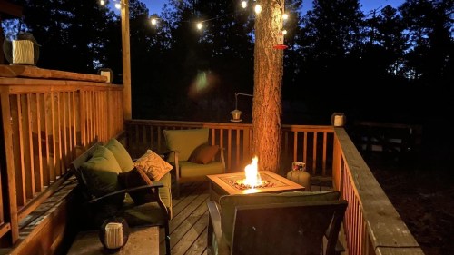 Cozy outdoor seating area with a fire pit, surrounded by trees and string lights at dusk.