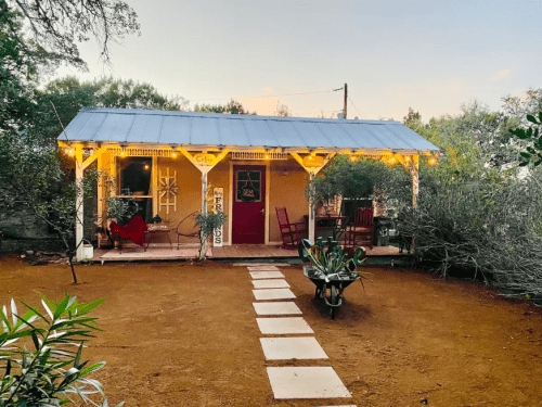 A cozy cottage with a porch, surrounded by greenery and a path leading to the entrance, illuminated by string lights.