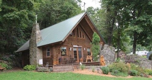 A cozy wooden cabin with a green metal roof, surrounded by trees and a stone pathway.