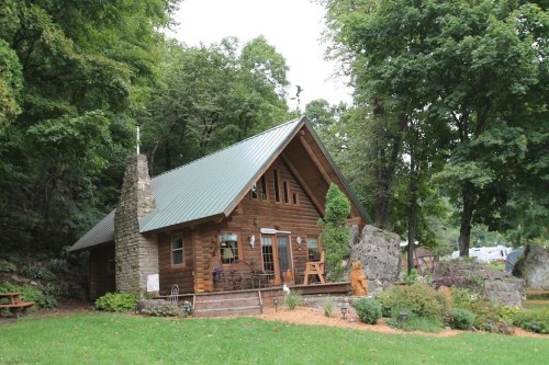 A charming wooden cabin with a green metal roof, surrounded by trees and a landscaped garden.