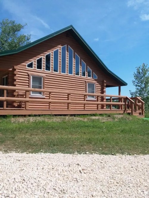 A log cabin with large windows and a green roof, surrounded by grass and trees under a clear blue sky.