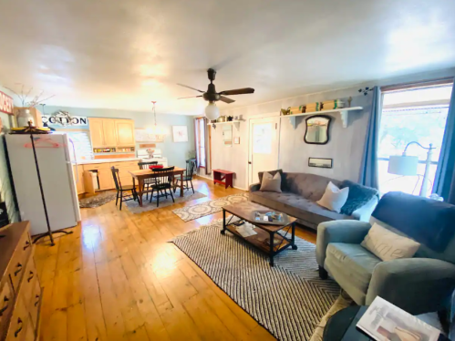 Cozy living room with a sofa, armchair, wooden floor, and a dining area visible in the background. Bright and inviting space.