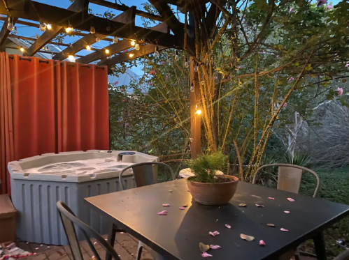 A cozy outdoor space with a hot tub, a table with petals, and string lights illuminating the area.