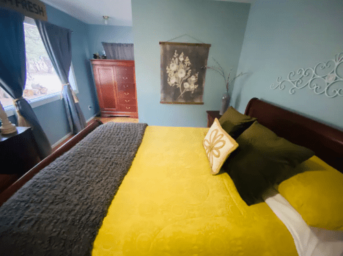 Cozy bedroom with a yellow bedspread, decorative pillows, and a window with curtains, featuring a dresser and wall art.