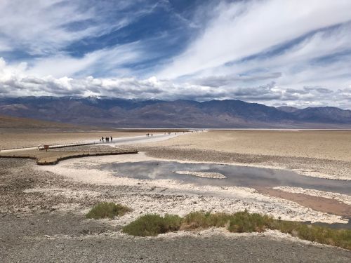 A wide view of a dry, cracked landscape with a wooden path and people walking, surrounded by mountains and cloudy skies.