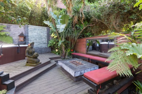 A serene outdoor space with a hot tub, stone statue, fire pit, and lush greenery surrounding wooden seating.