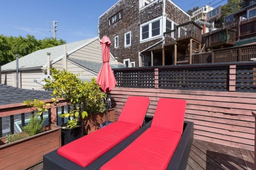 Two red lounge chairs on a wooden deck, with a red umbrella and a view of nearby houses and greenery.