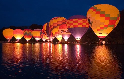 Colorful hot air balloons illuminated at night, reflecting on a calm lake with a dark sky in the background.