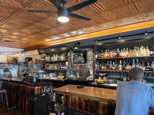 A cozy bar interior featuring a wooden counter, shelves of liquor, and a decorative ceiling. A person sits at the bar.
