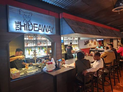 A lively bar scene at The Hideaway, with patrons seated at the counter and a bartender preparing drinks.