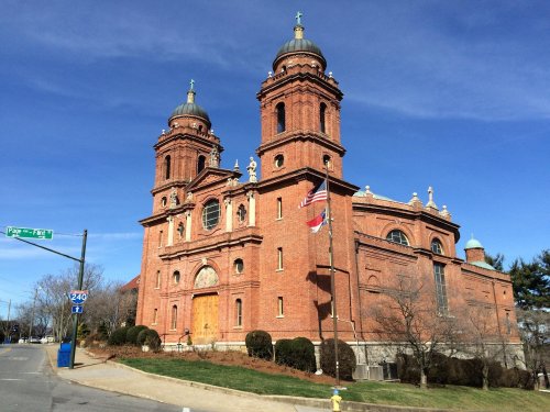 Historic brick church with twin towers, surrounded by greenery and a flag, under a clear blue sky.