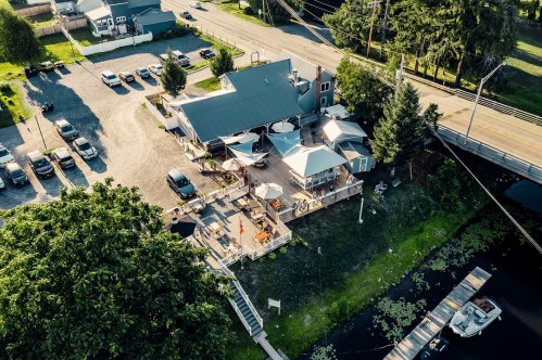 Aerial view of a riverside restaurant with outdoor seating, surrounded by trees and parked cars.