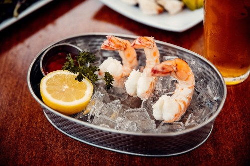 A metal bowl filled with ice, featuring shrimp, a lemon slice, and dipping sauce, with a drink in the background.