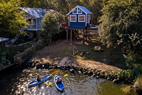 Aerial view of a blue house on stilts by a river, with two kayakers paddling nearby and lush greenery surrounding the area.