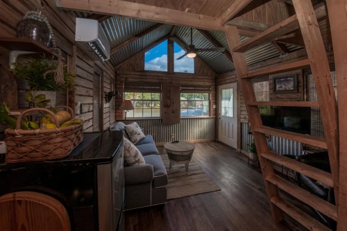 Cozy interior of a rustic cabin with wooden walls, a sofa, and a lofted sleeping area. Natural light from large windows.
