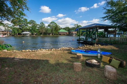 A serene lakeside scene with a kayak, seating area, and a dock under a blue sky with scattered clouds.