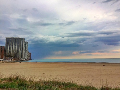 A sandy beach with a calm lake, cloudy skies, and tall buildings in the background.