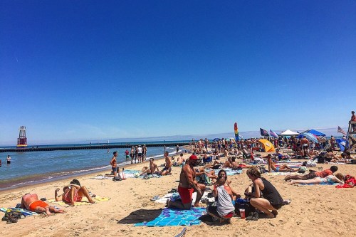 A busy beach scene with people sunbathing, umbrellas, and a pier in the background under a clear blue sky.