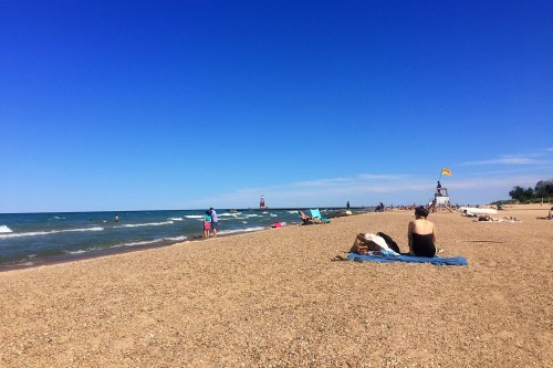 A sunny beach scene with people relaxing on the sand, gentle waves, and a distant lighthouse under a clear blue sky.