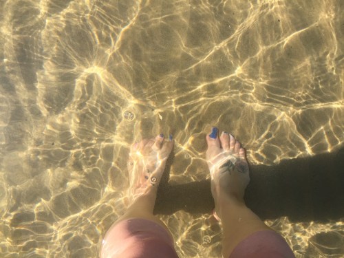 Feet with blue nail polish submerged in clear water, with sunlight creating rippling reflections on the sandy bottom.