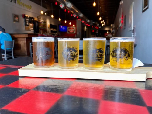 A flight of four craft beers on a wooden paddle, set on a checkered table in a brewery.