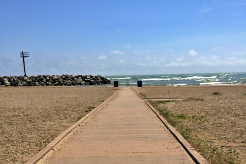 A wooden walkway leads to the ocean, with rocky shore and blue sky in the background. Trash bins are visible near the water.
