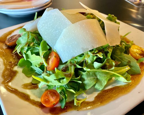 A fresh salad with mixed greens, cherry tomatoes, and shaved cheese drizzled with dressing on a white plate.
