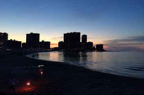 Silhouetted buildings along a beach at dusk, with a calm lake and a few people holding sparklers.