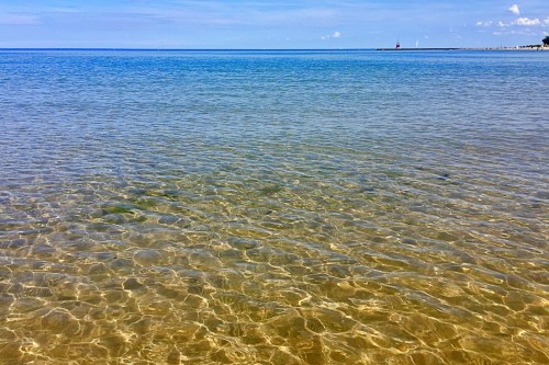 Clear, calm water with gentle ripples, reflecting a blue sky and distant shoreline under a sunny day.