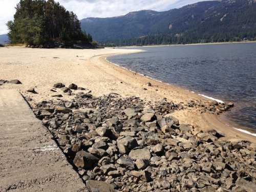 A sandy beach curves along a calm lake, with rocky areas and trees in the background under a partly cloudy sky.