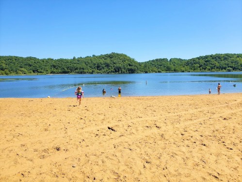 A sandy beach by a calm lake, with people enjoying the water and green hills in the background under a clear blue sky.