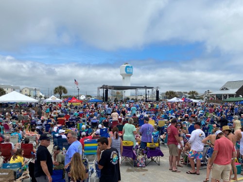 A large crowd gathers at a beach event, with tents and a stage in the background under a cloudy sky.